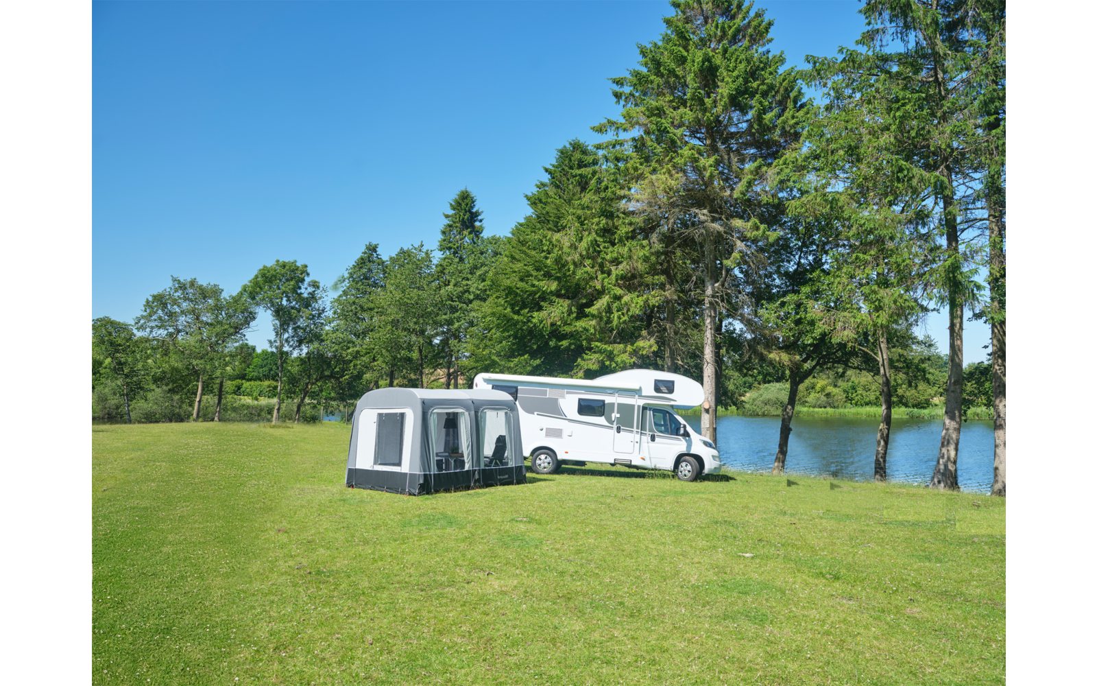 Weißes Wohnmobil mit grauem Vorzelt auf grüner Wiese neben einem See, im Hintergrund Bäume und blauer Himmel