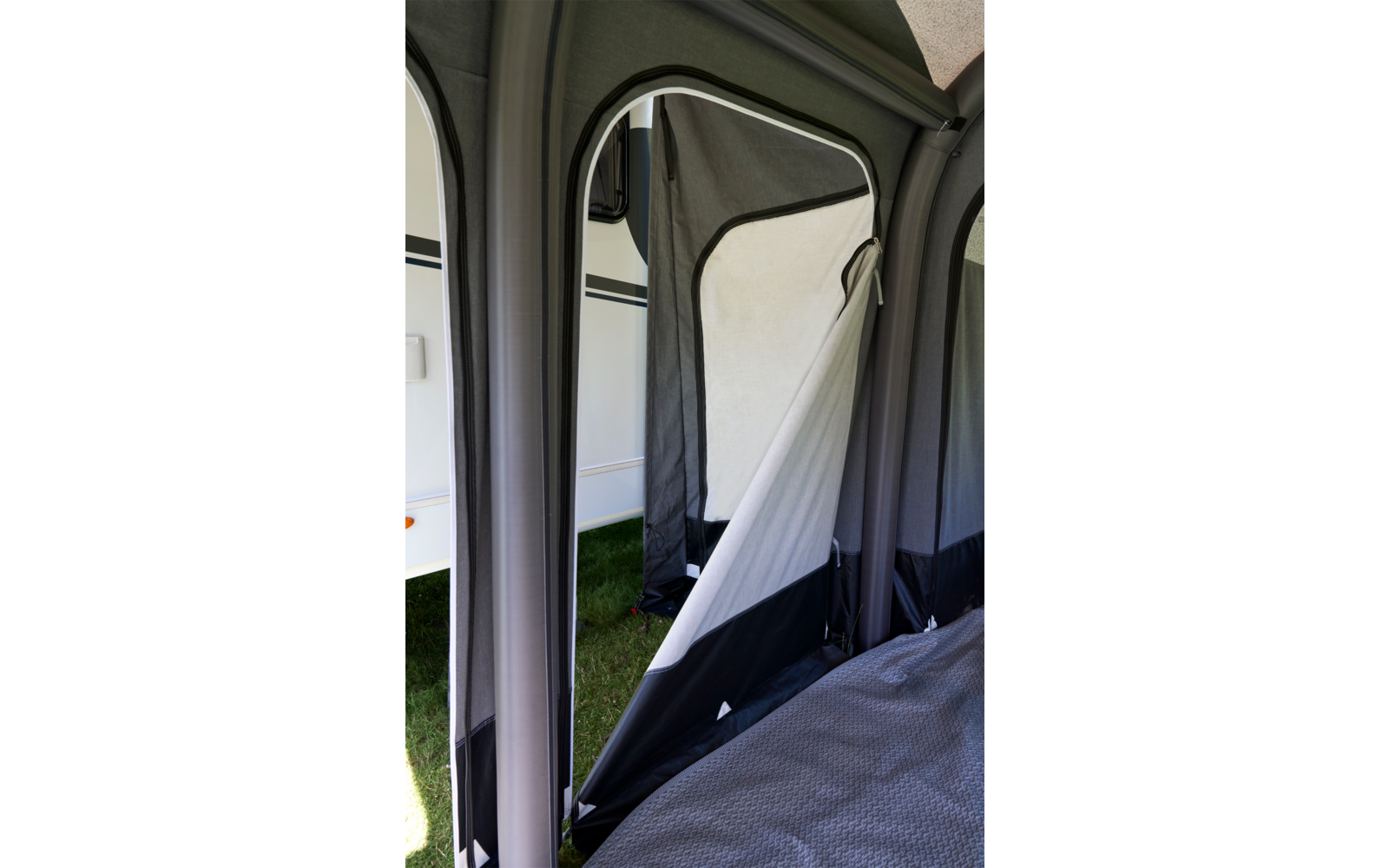 Innenseite eines grauen Zeltes mit offenem Seiteneingang; durch die Öffnung ist die weiße Wand eines Wohnwagens und Gras sichtbar; innen eine Matratze mit grauem Bezug