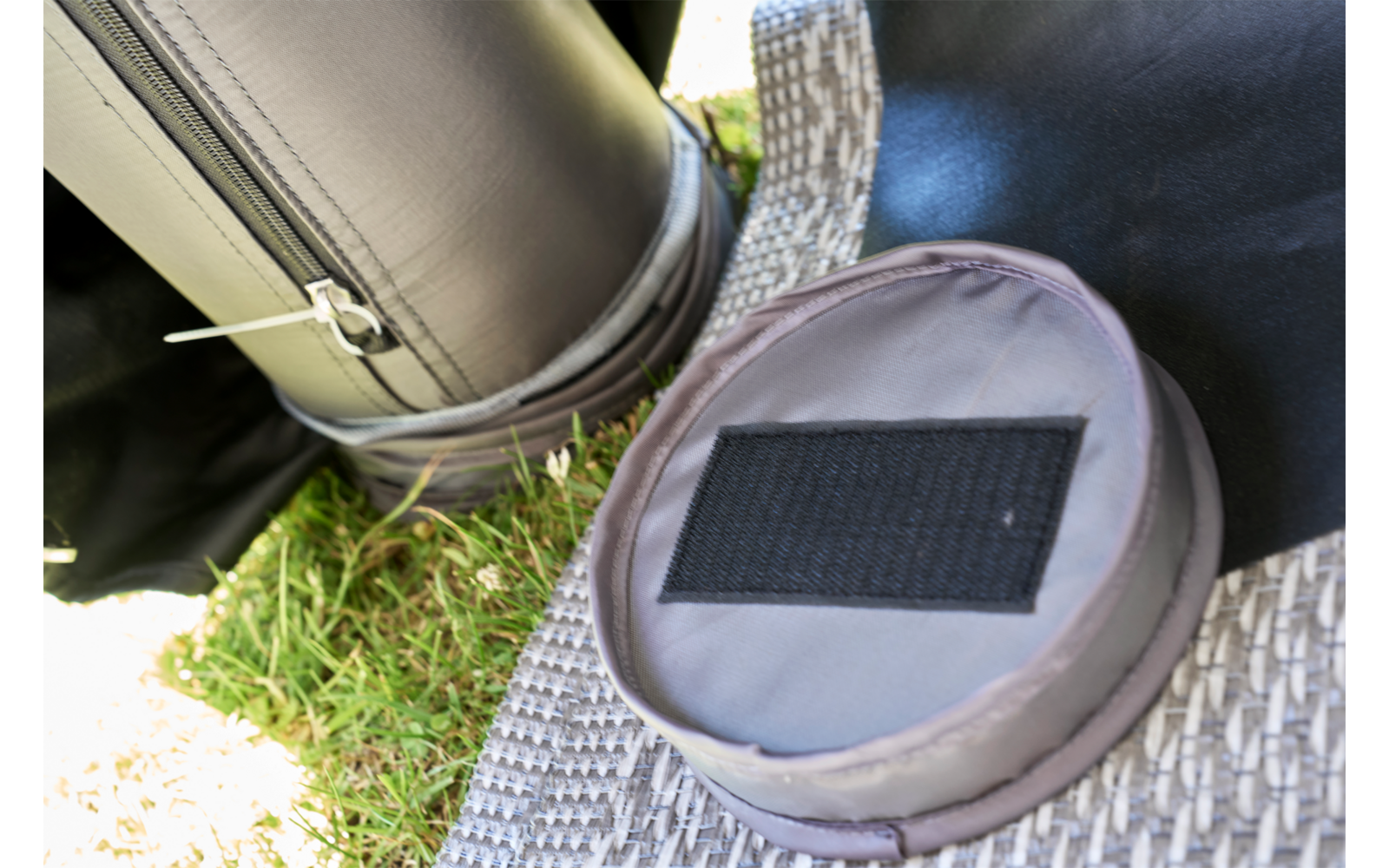 Runder grauer Stoffbeutel mit rechteckigem schwarzem Klettfeld auf geflochtener Matte, daneben aufgerolltes schwarzes Polster und Gras