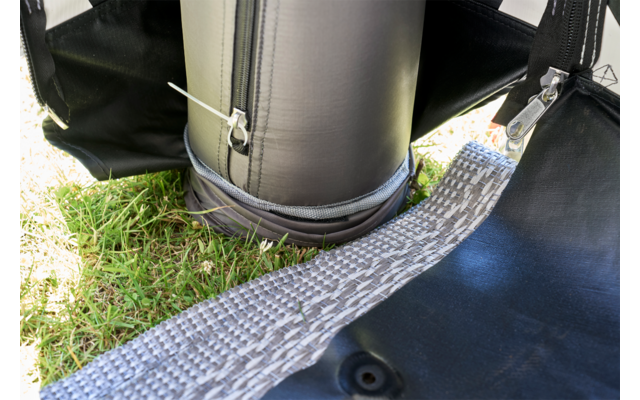 Schwarzer zylindrischer Gegenstand mit Reißverschluss und weißem Kabelbinder, grauer gewebter Gurt auf Gras, schwarze Plane mit Metallhaken