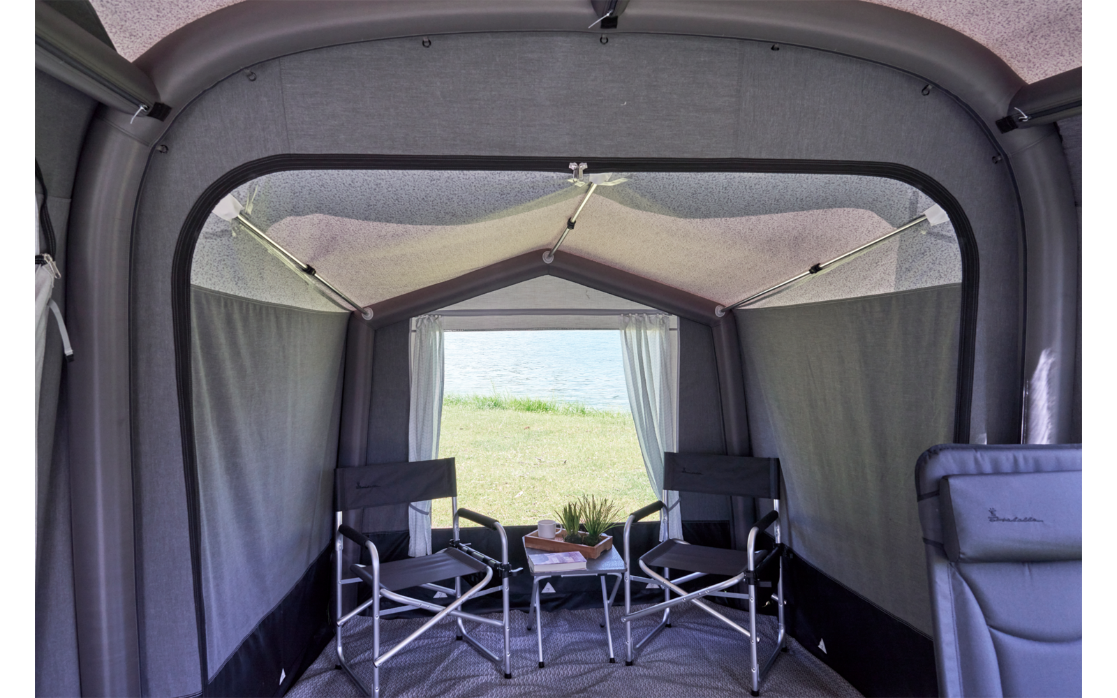 Innenraum eines grauen Vorzelts mit zwei klappbaren Campingstühlen und einem kleinen Tisch mit einer Topfpflanze und Gegenständen, vorderer Eingang geöffnet, im Hintergrund Gras und Wasser