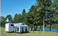 Caravane avec auvent sur une prairie au bord d'un lac, deux personnes debout devant, arbres et ciel bleu en arrière-plan