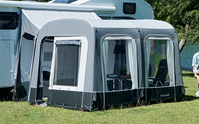Caravana con toldo en un campo de hierba junto a un lago, dos personas de pie delante, árboles y cielo azul al fondo