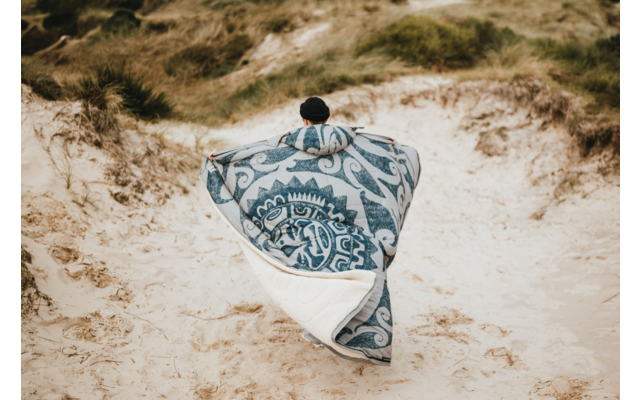 Personne avec bonnet noir, vue de dos sur des dunes de sable, enveloppée dans une grande couverture à motif bleu et blanc