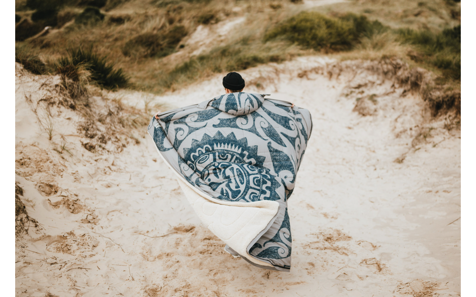 Personne avec bonnet noir, vue de dos sur des dunes de sable, enveloppée dans une grande couverture à motif bleu et blanc