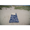 Serviette de plage bleue à motifs avec une petite serviette blanche en haut, posée sur le sable ; dunes herbeuses, deux poteaux en bois et ciel bleu en arrière-plan