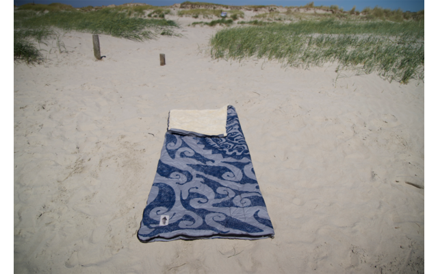 Serviette de plage bleue à motifs avec une petite serviette blanche en haut, posée sur le sable ; dunes herbeuses, deux poteaux en bois et ciel bleu en arrière-plan