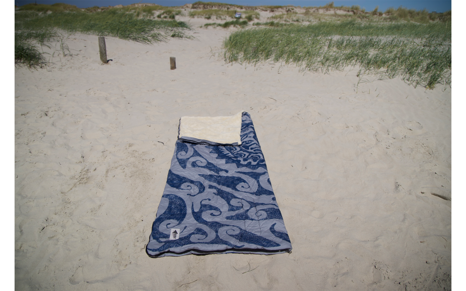 Serviette de plage bleue à motifs avec une petite serviette blanche en haut, posée sur le sable ; dunes herbeuses, deux poteaux en bois et ciel bleu en arrière-plan