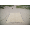 Couverture de plage rectangulaire beige avec bord sombre sur du sable clair, dunes avec herbe de plage et deux poteaux en bois en arrière‑plan