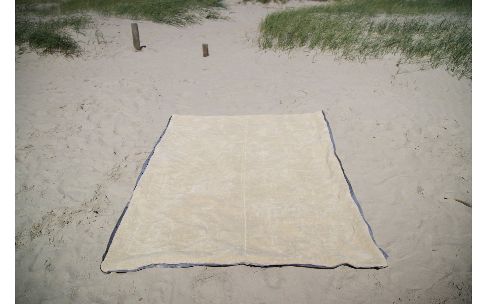 Couverture de plage rectangulaire beige avec bord sombre sur du sable clair, dunes avec herbe de plage et deux poteaux en bois en arrière‑plan