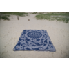Serviette rectangulaire bleue avec motif ornemental blanc posée sur du sable clair; herbe de dune et deux poteaux en bois en arrière-plan