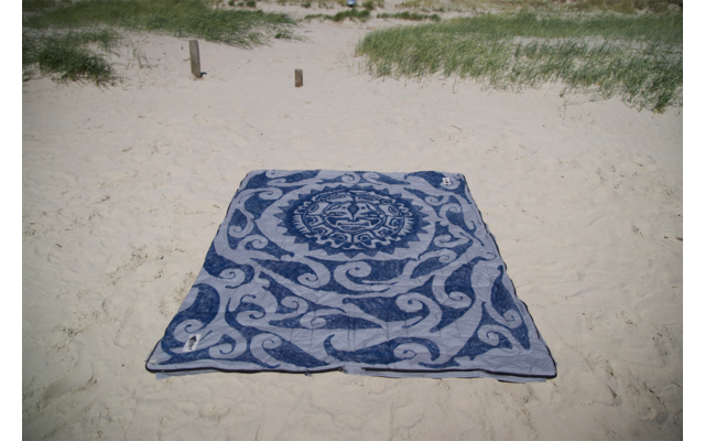 Serviette rectangulaire bleue avec motif ornemental blanc posée sur du sable clair; herbe de dune et deux poteaux en bois en arrière-plan