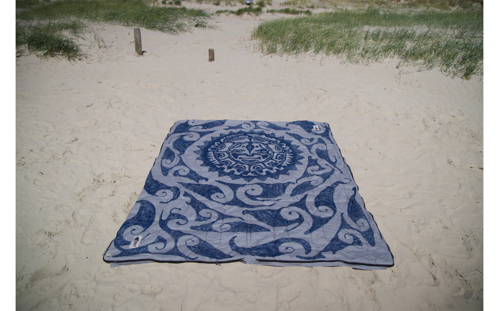 Serviette rectangulaire bleue avec motif ornemental blanc posée sur du sable clair; herbe de dune et deux poteaux en bois en arrière-plan