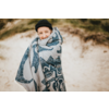 Personne portant un bonnet noir, enveloppée dans une couverture à motifs, devant des dunes de sable, souriant à l'appareil photo