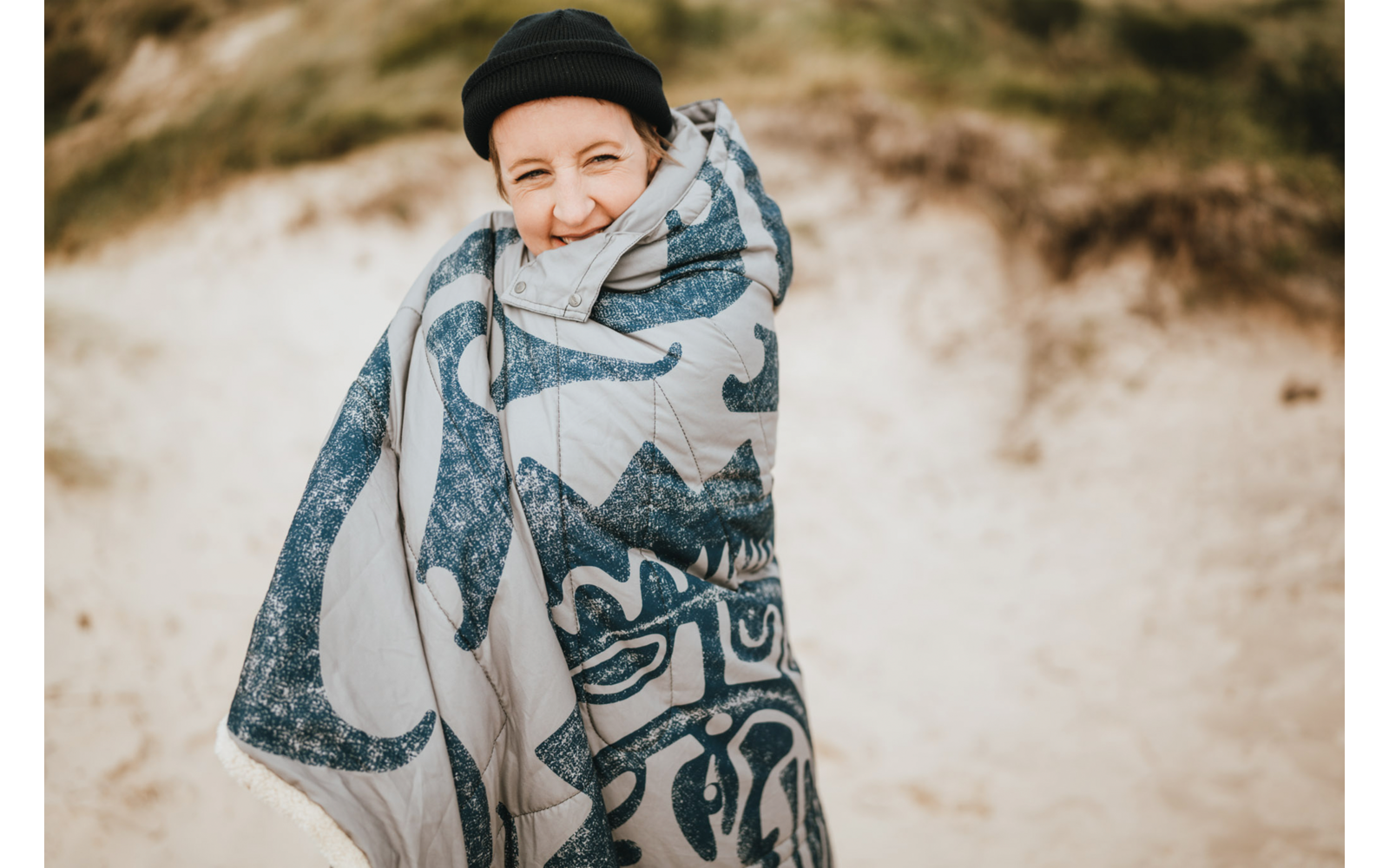 Personne portant un bonnet noir, enveloppée dans une couverture à motifs, devant des dunes de sable, souriant à l'appareil photo