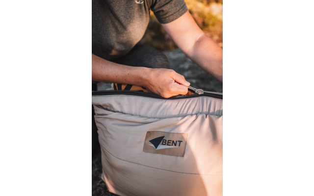 Mains tirant une fermeture éclair d'un sac matelassé beige clair avec un écusson 'BENT' ; personne portant un T-shirt gris foncé à manches courtes ; arrière-plan flou d'herbe
