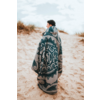 Personne debout sur une dune de sable, enveloppée d'une grande couverture à motifs, portant un bonnet noir, regardant vers la gauche ; herbes de dune et ciel nuageux en arrière-plan