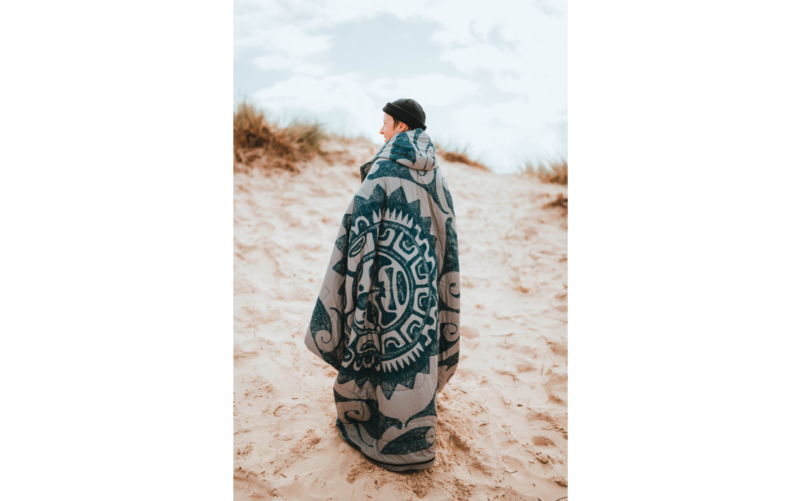 Personne debout sur une dune de sable, enveloppée d'une grande couverture à motifs, portant un bonnet noir, regardant vers la gauche ; herbes de dune et ciel nuageux en arrière-plan
