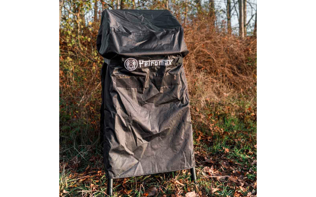 Schwarze Abdeckhaube mit Petromax-Logo über einem Gegenstand; zwei Metallbeine sichtbar; Boden mit trockenen Blättern und kahlen Sträuchern