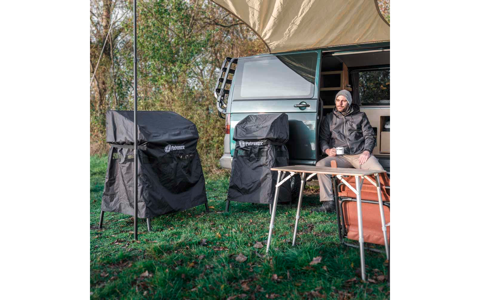 Person sitzt auf Klappstuhl neben offenem Van unter Markise, hält Tasse; zwei schwarze, abgedeckte Geräte auf Ständern, orange Tasche, Gras und Bäume im Hintergrund