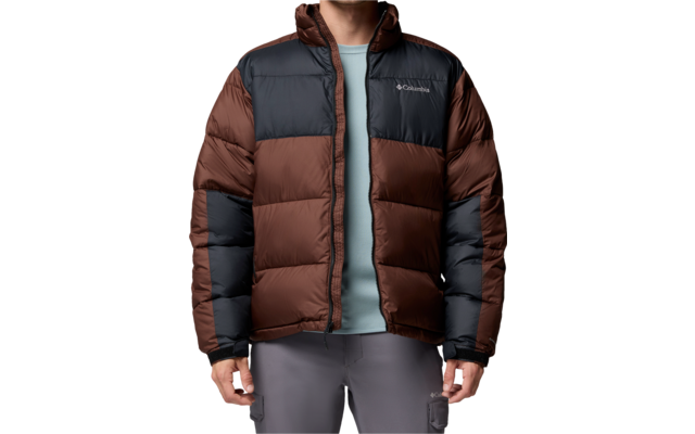 Stehende Person mit kurzen braunen Haaren, braun-schwarze gesteppte Daunenjacke, hellblaues T‑Shirt, graue Hose, weißer Hintergrund
