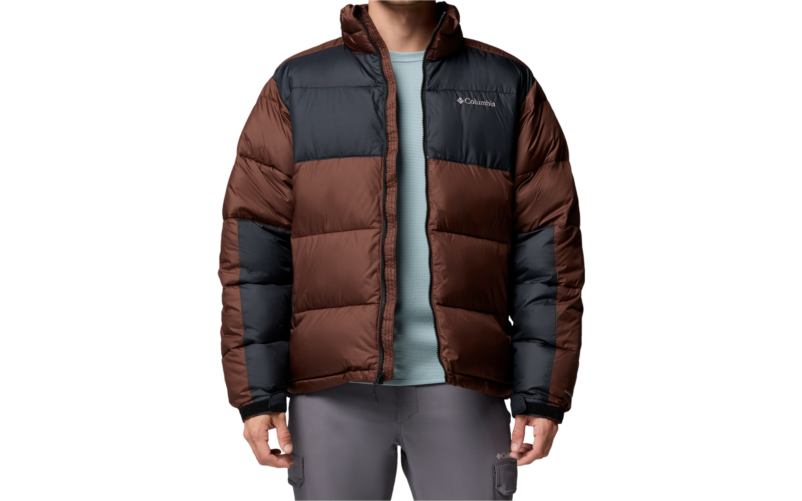 Stehende Person mit kurzen braunen Haaren, braun-schwarze gesteppte Daunenjacke, hellblaues T‑Shirt, graue Hose, weißer Hintergrund
