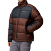 Person mit kurzem dunklem Haar, trägt eine braun-dunkelgraue Daunenjacke mit Columbia-Logo, Hände in den Jackentaschen, hellgraue Hose, weißer Hintergrund