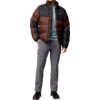 Ein stehender Mann in gestepptem braun-dunkelgrauem Steppjacke, grauem Pullover, grauen Hosen und schwarzen Schuhen, Blick nach rechts, eine Hand in der Jackentasche