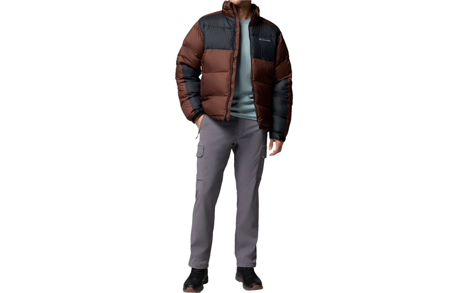 Ein stehender Mann in gestepptem braun-dunkelgrauem Steppjacke, grauem Pullover, grauen Hosen und schwarzen Schuhen, Blick nach rechts, eine Hand in der Jackentasche