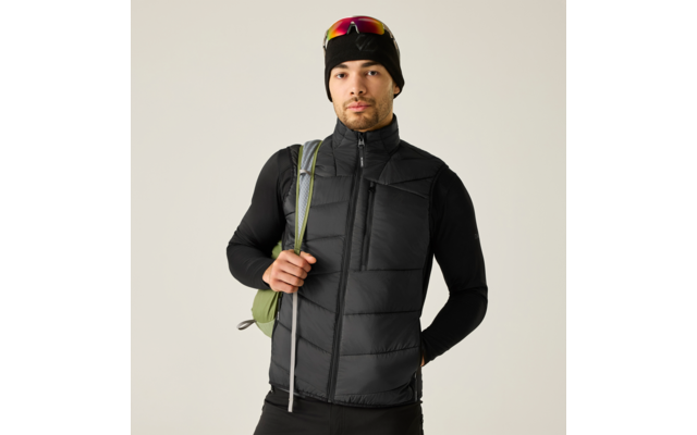 Person in schwarzer Steppweste und schwarzem Langarmshirt, schwarze Mütze mit aufgesetzter Sonnenbrille, hält grünen Rucksackriemen, heller Hintergrund