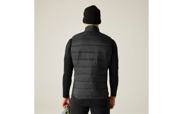 Person von hinten, trägt schwarze gesteppte Weste, schwarzes Langarmshirt, schwarze Mütze und schwarze Hose, hält eine grüne Jacke in der linken Hand; heller einfarbiger Hintergrund