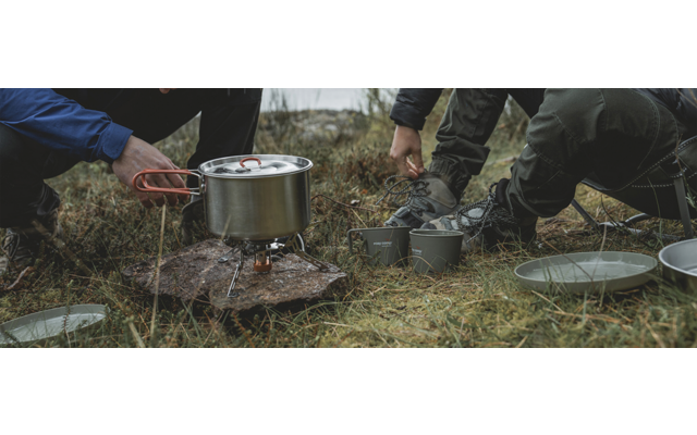 Hände bedienen einen Campingkocher mit einem Kochtopf darauf; Tassen und Teller liegen auf dem Gras