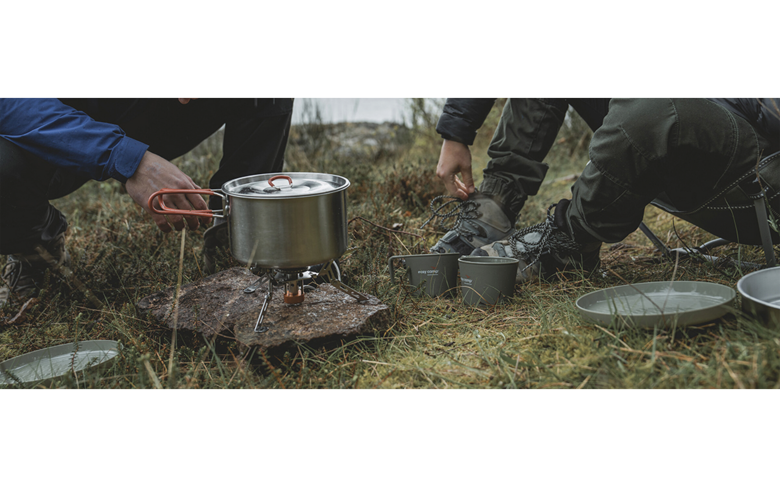 Hände bedienen einen Campingkocher mit einem Kochtopf darauf; Tassen und Teller liegen auf dem Gras
