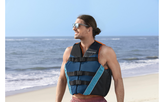 Person mit zusammengebundenem Haar, Sonnenbrille und blau-schwarzer Rettungsweste steht auf einem Sandstrand, Blick nach rechts, rote Badeshorts, Meer mit Wellen im Hintergrund