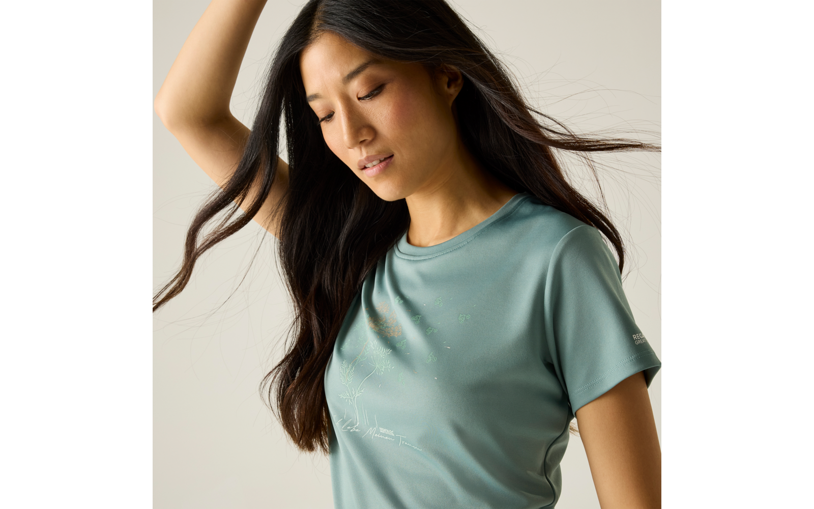 Personne aux longs cheveux foncés regardant vers le bas, portant un T‑shirt vert menthe à manches courtes avec un petit motif sur la poitrine, un bras levé, fond beige uni