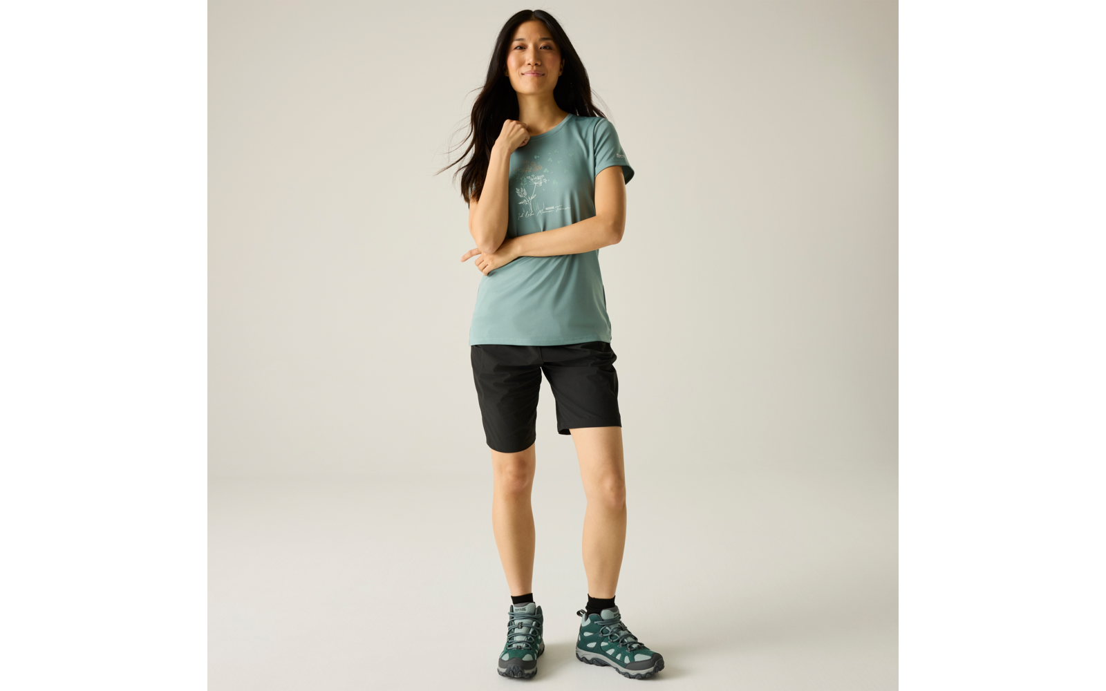 Femme debout aux longs cheveux foncés, T‑shirt turquoise avec petite impression blanche, short noir au‑dessus du genou, chaussures de randonnée vertes, main droite au niveau du cou, bras gauche devant le corps, fond uni clair