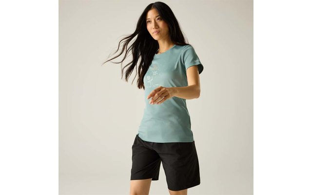 Personne aux longs cheveux foncés portant un t-shirt bleu-vert avec un petit motif et un short noir jusqu'aux genoux, marchant devant un fond clair