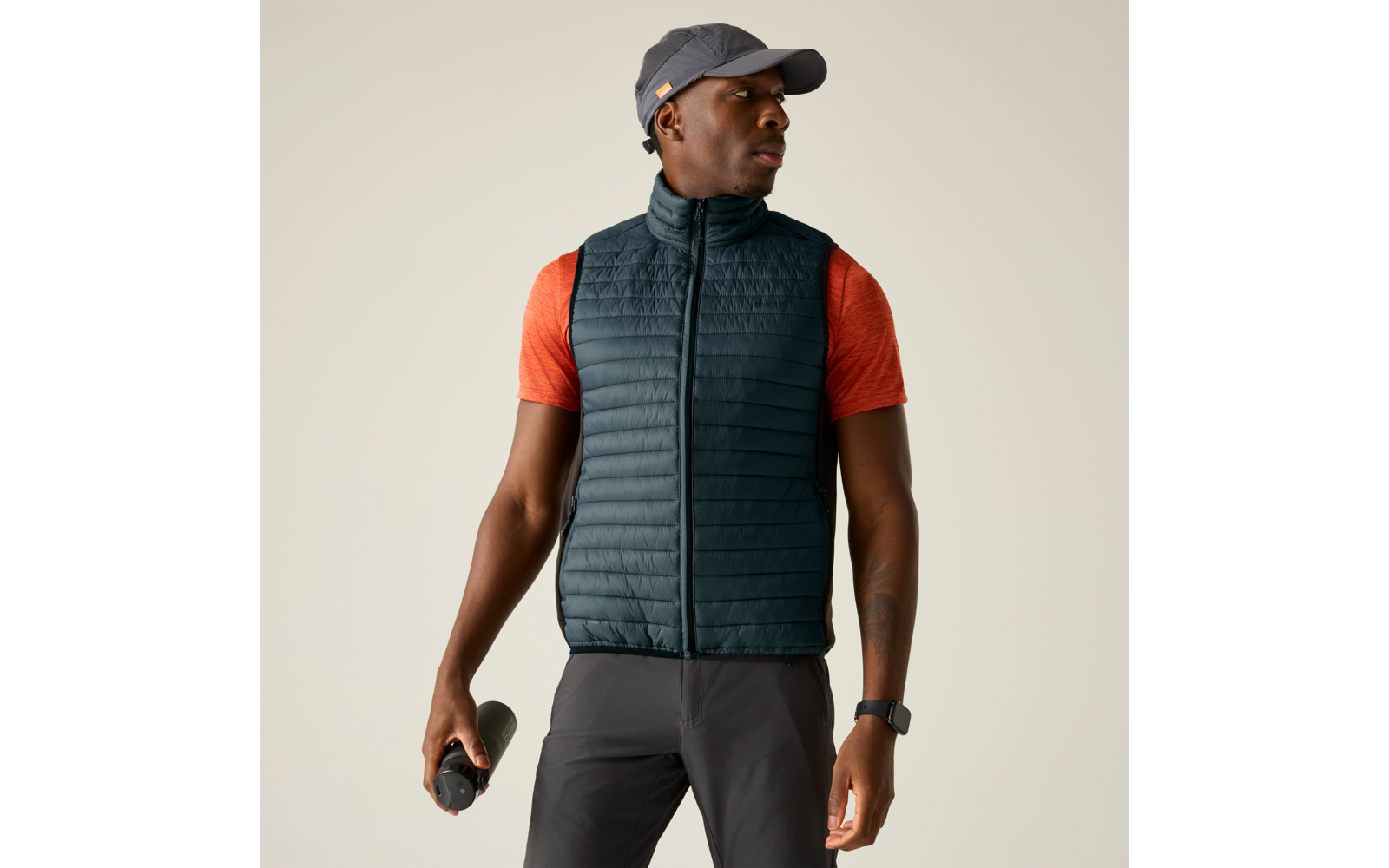 Person mit grauer Kappe, orangefarbenem Kurzarmshirt und dunkler gesteppter Weste, hält eine Trinkflasche, trägt eine Uhr am linken Handgelenk, Blick zur Seite, heller einfarbiger Hintergrund