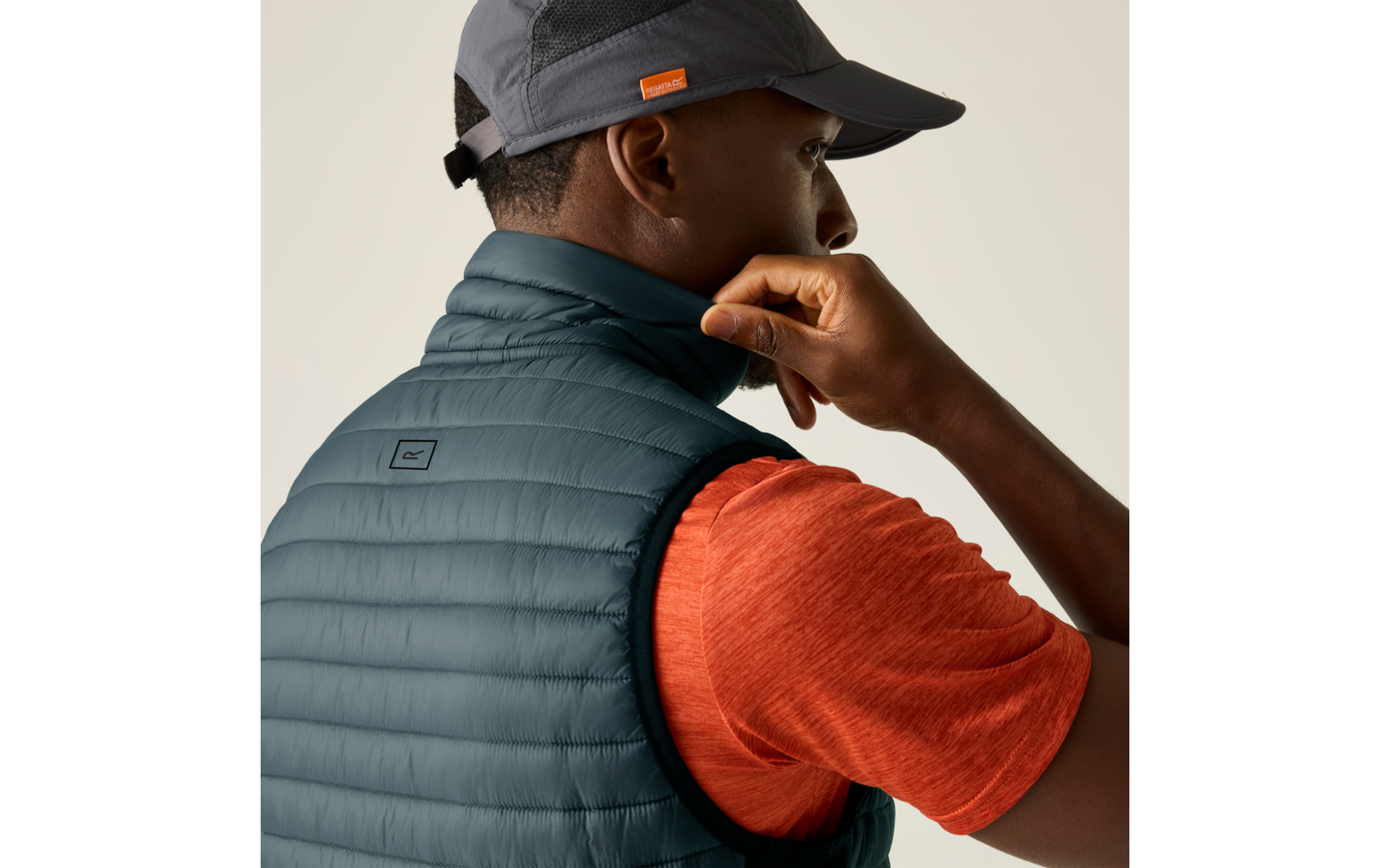 Person von hinten und im Profil, trägt graue Kappe, dunkelblaue gesteppte ärmellose Weste über orangefarbenem T‑Shirt, rechte Hand am Kragen, heller einfarbiger Hintergrund