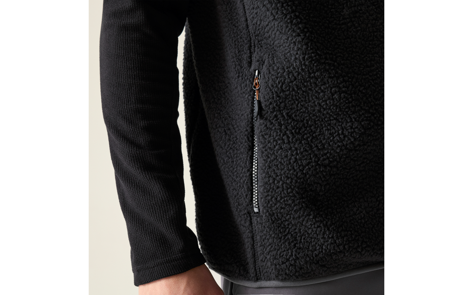Nahaufnahme eines Oberkörpers in schwarzem Teddy-Fleece mit seitlicher Reißverschlusstasche, schwarzem Langarmshirt darunter, sichtbare Hand und graue Hose vor hellem Hintergrund