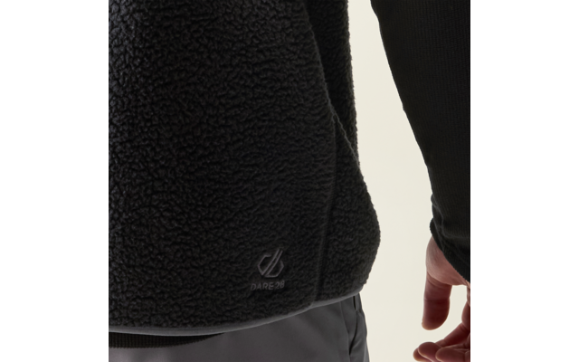 Teilansicht einer schwarzen, strukturierten Fleecejacke mit sichtbarem 'Dare 2b'-Logo am Saum, angrenzende graue Hose, sichtbare Hand und Unterarm mit schwarzem Ärmelsaum, heller Hintergrund