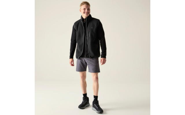 Eine stehende Person mit kurzen blonden Locken, lächelnd, trägt eine schwarze Fleeceweste über einem schwarzen Langarmshirt, graue Shorts, schwarze Socken und schwarze Sportschuhe vor hellem Hintergrund