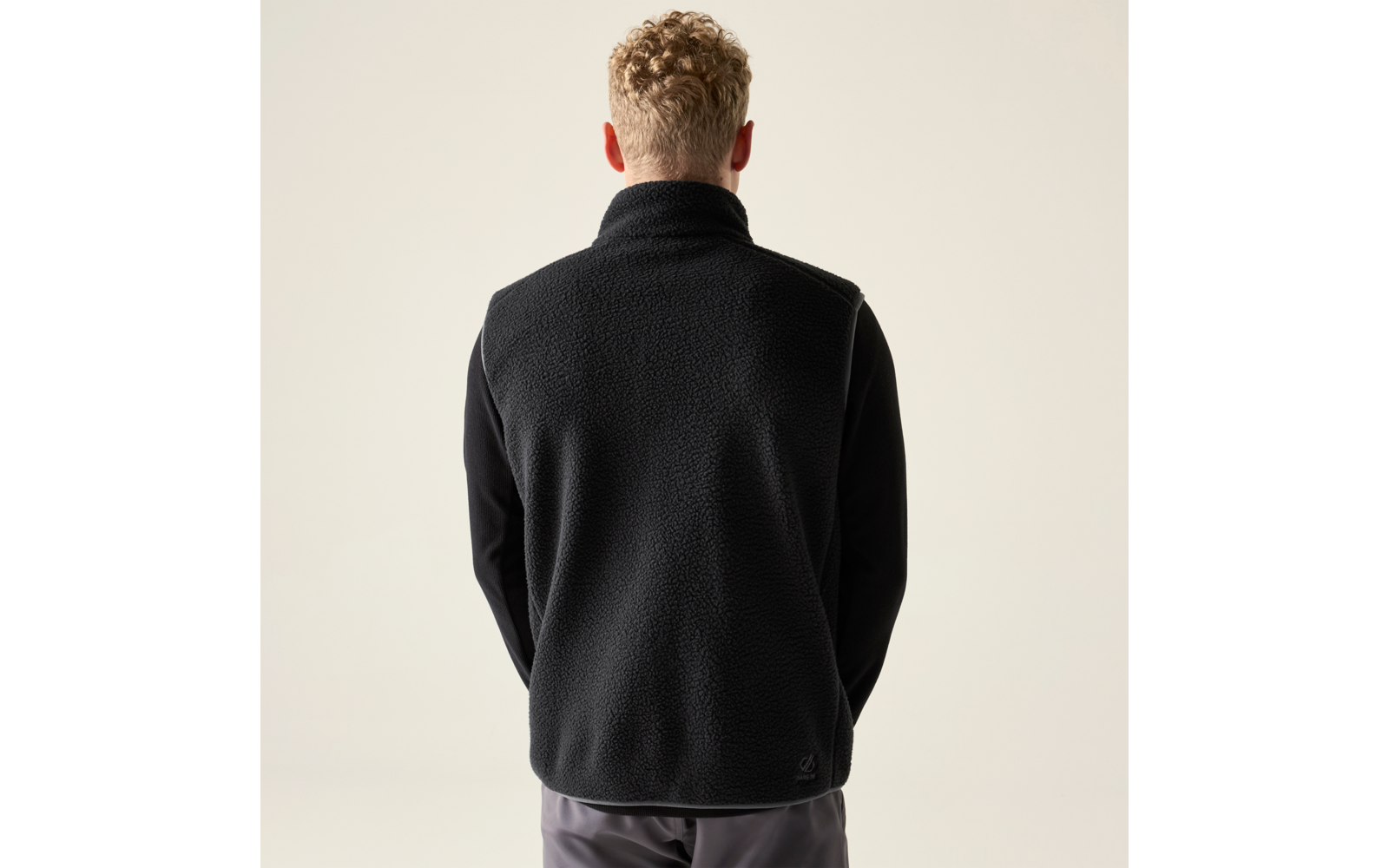 Rückansicht einer Person mit kurzen lockigen blonden Haaren, die eine schwarze Fleeceweste über einem schwarzen Langarmshirt trägt