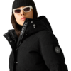 Person mit weißer Sonnenbrille, schwarzer Strickmütze und schwarzer gesteppter Jacke mit Kapuze, Reißverschluss mit Schriftzug 'DARE2B' und rundem Logo-Patch auf dem Ärmel; Gesicht teilweise sichtbar