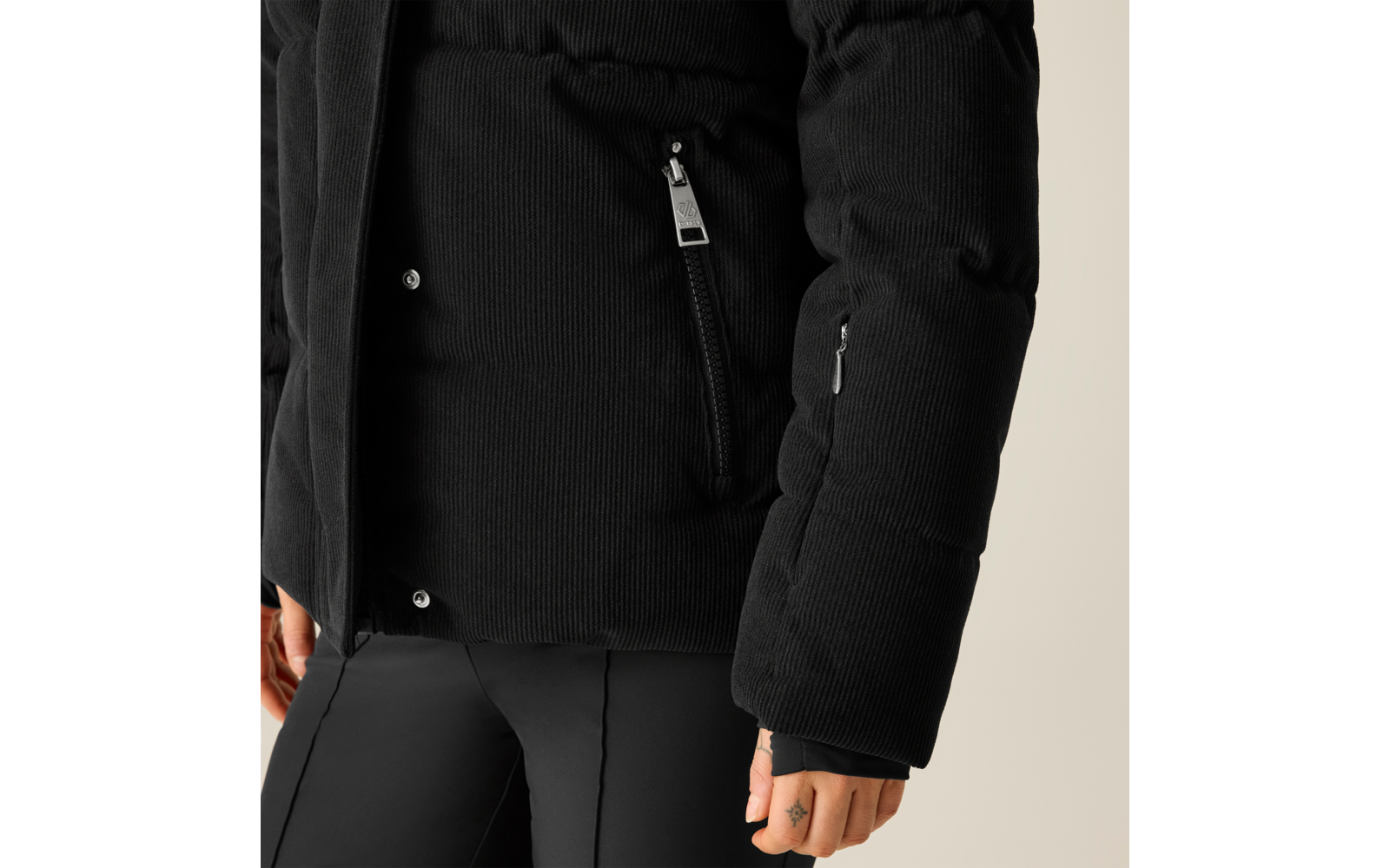 Schwarze gesteppte Cordjacke mit Reißverschlusstasche, schwarze Hose, sichtbare Hand mit kleinem Fingertattoo