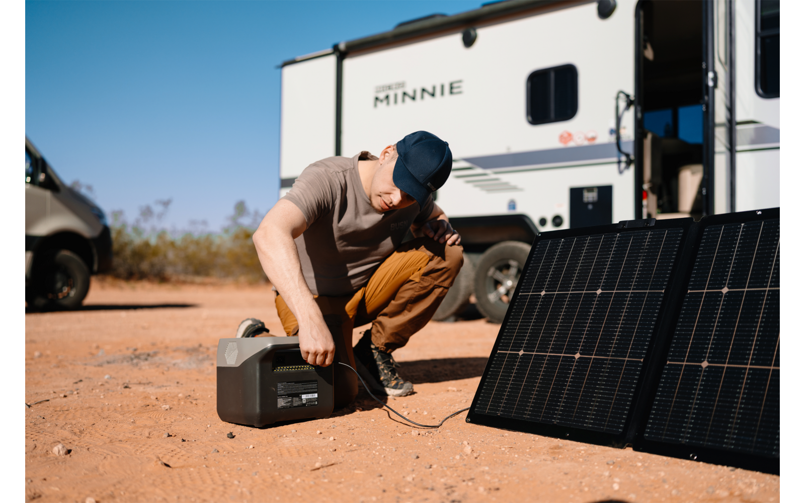 Mann kniet auf Boden vor Solarpanel und öffnet Werkzeugkoffer, im Hintergrund Wohnmobil