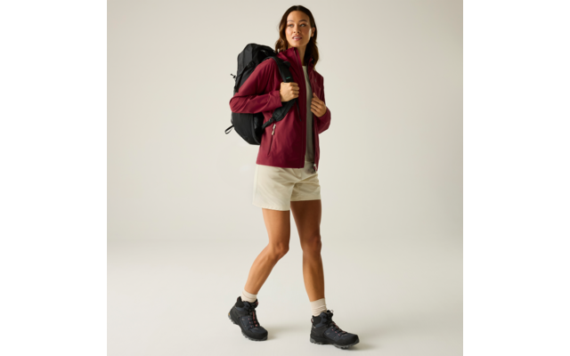 Vrouw staat voor een lichte achtergrond, draagt een bordeauxrode jas, beige korte broek, witte sokken, zwarte wandelschoenen en een zwarte rugzak