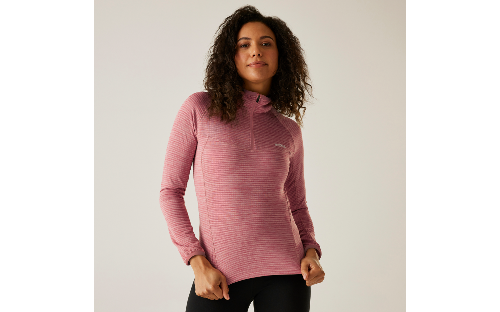 Frau mit lockigem Haar trägt einen rosa Pullover und steht vor weißer Wand