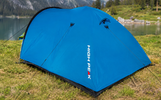Blaues Kuppelzelt mit Aufschrift "HIGH PEAK" auf Gras am Ufer eines Sees, im Hintergrund Berge und Bäume