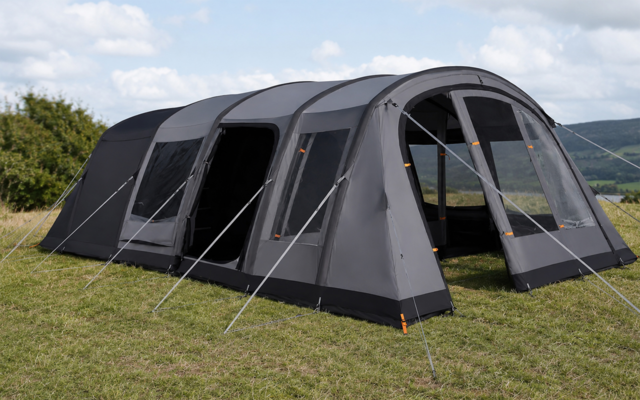 Grande tenda a tunnel grigia con più archi, ingresso aperto e tiranti, su prato; colline e cielo nuvoloso sullo sfondo